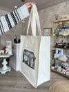 White tote bag with black design held by a person in a room with decor elements.